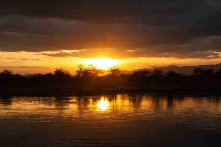 Sunset over a calm river with the sun's reflection on the water, silhouetted trees against a vibrant orange sky, and distant mountains.